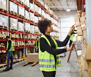 Female warehouse worker stacking boxes onto the rack. Woman working in large distribution warehouse. she is wearing uniform and reflective clothing, with colleagues working in background.