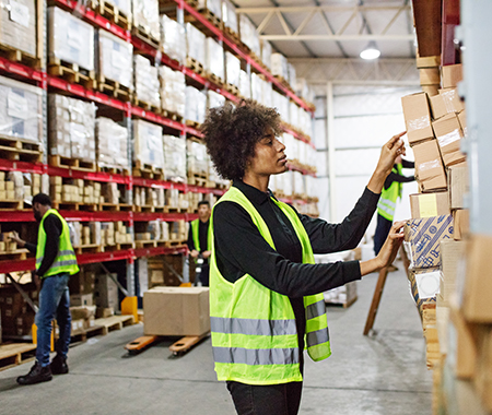 Female warehouse worker stacking boxes onto the rack. Woman working in large distribution warehouse. she is wearing uniform and reflective clothing, with colleagues working in background.