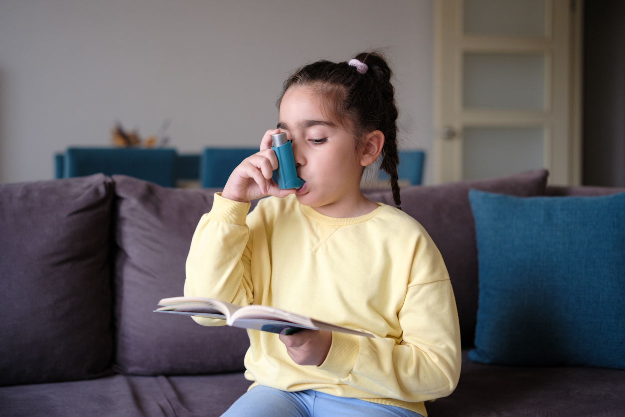 Little girl uses an asthma inhaler while reading book at home