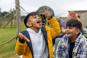 Front view of two young brothers at an outdoor adventure playground. They are sitting on a wooden bench, one boy is holding string cheese up in the air with his head back and his mouth wide open.