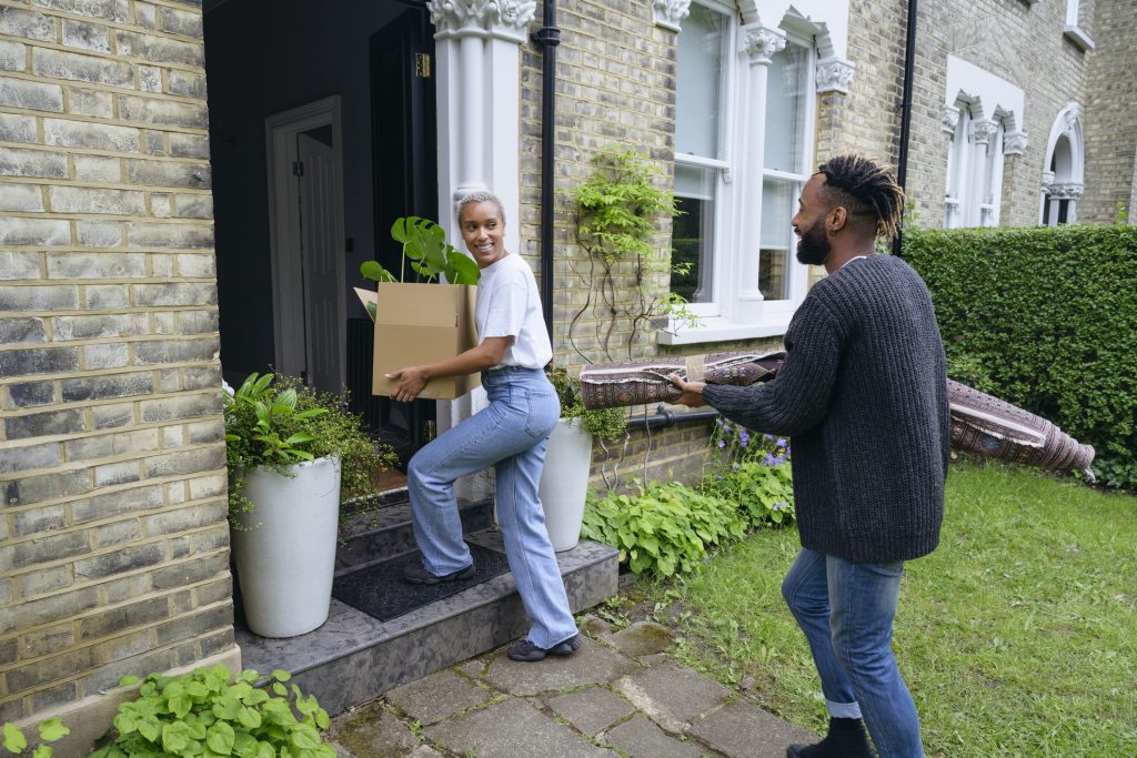 Full length view of man and woman in casual clothing smiling at each other as they carry cardboard box and rug up front steps on moving day.